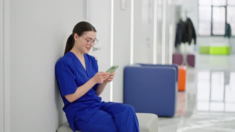 Confident-and-happy-girl-brunette-doctor-with-glasses-in-a-blue-uniform-sits-on-the-side-in-the-corridor-of-a-modern-clinic-and-types-on-a-green-smartphone