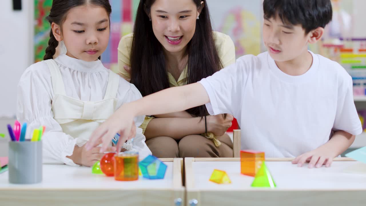 明るい教室で、笑顔の教師が2人の小学生がカラフルな幾何学的立体を探索するのを手伝っています。