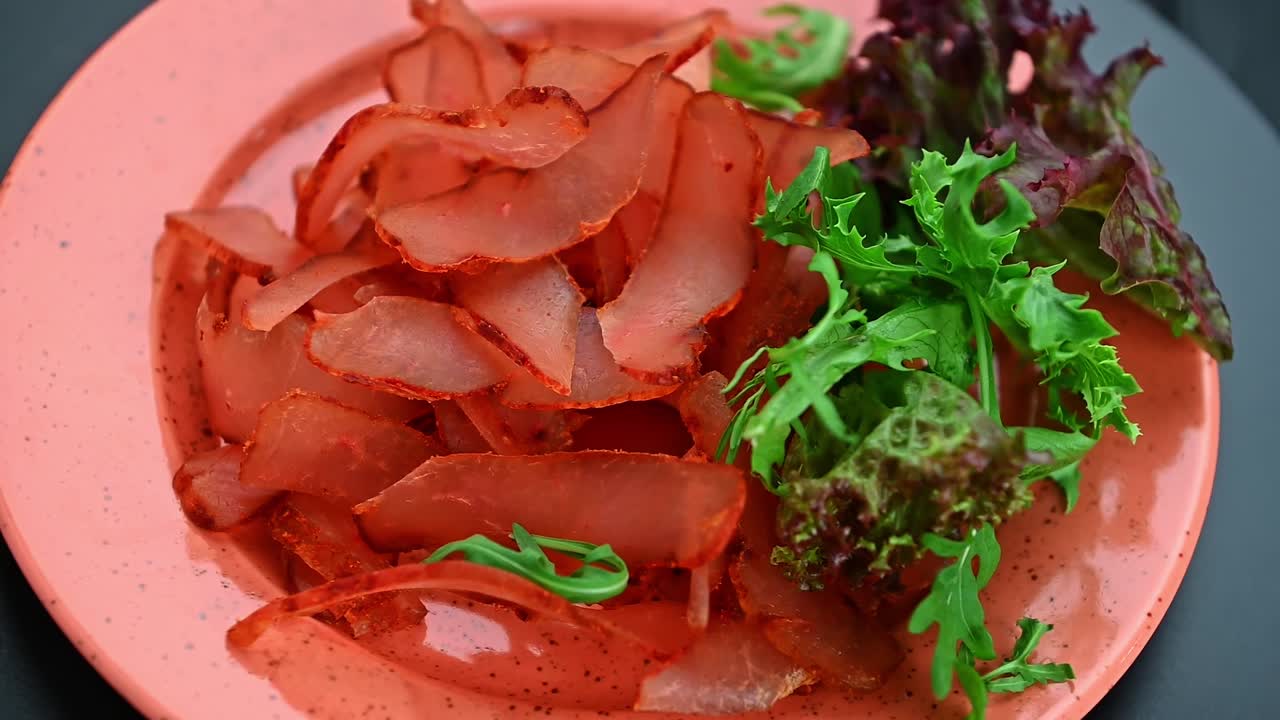 Fresh slices of meat arranged on a pink plate with herbs on the side. The dish shows thin slices of meat on a pink plate with green herbs and salad