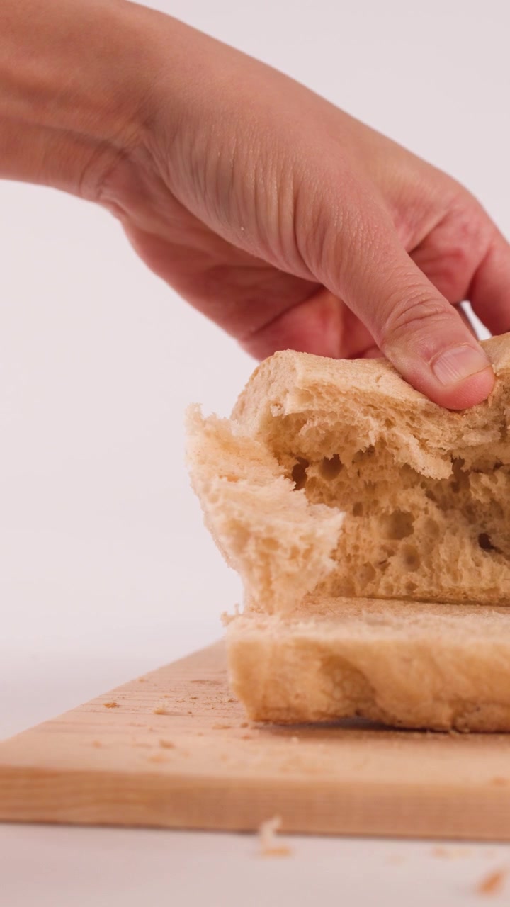 Close-up of hands pulling apart soft focaccia bread on a wooden board against white background.