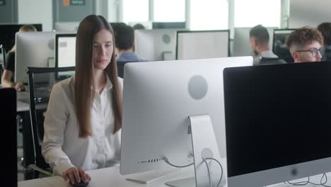 joven atractiva trabajando en una computadora de cubierta mientras trabaja en una gran oficina de espacio abierto. retrato de mujer de negocios positiva mirando la pantalla de la computadora en el interior