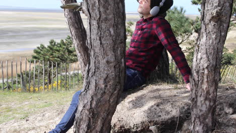 shot of an authentic hipster man listening to music with headphones whilst sitting in the sun against a tree trunk