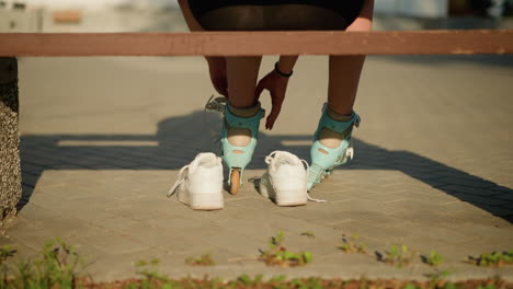 back view of individual adjusting straps on cyan roller skates of left leg while sitting on bench outdoors, with black bangle on right hand, white sneakers placed behind