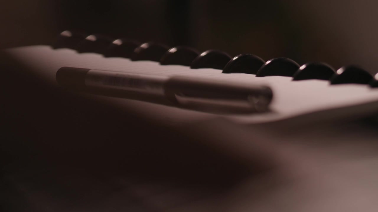Dita che scrivono su una tastiera in primo piano sfocato con una penna e un blocco note a fuoco sullo sfondo.