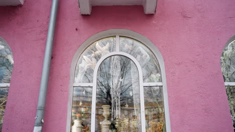 close-up view of a pink-colored house, featuring decorative windows with intricate designs, with flower vessel seen through the window glass