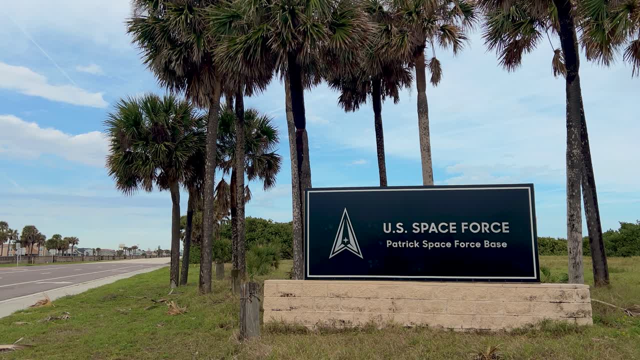 Wide static shot of U.S. Space Force marquee sign, centered, on a sunny Florida day. Quiet road on left, palm trees sway, vibrant coastal scene, clear sky, warm summer vibe, military base, Cocoa Beach