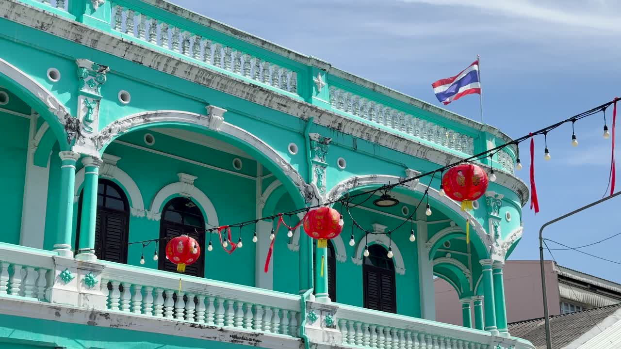 Turquoise colonial building with red lanterns, Thai flags, daylight, and slight upward camera movement