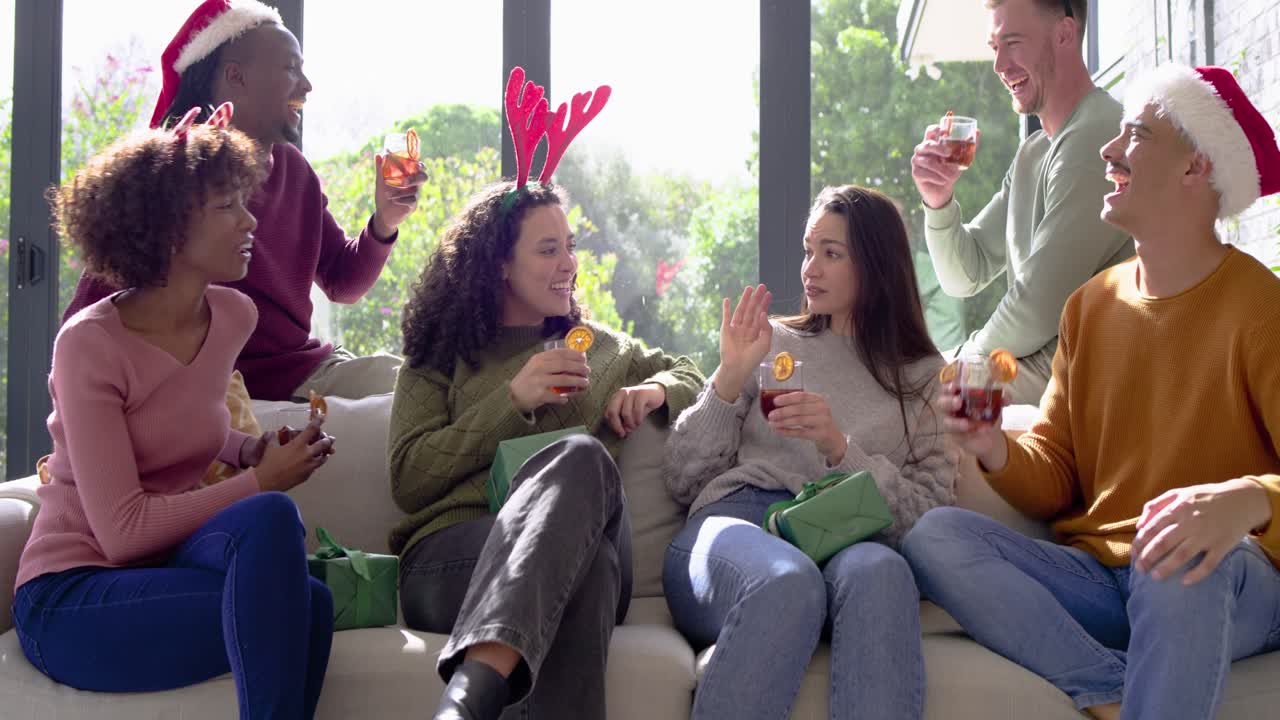 Diverse friends raising holiday cocktails on light sofa at home, clinking glasses and laughing