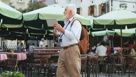 senior man walking as a tourist at a cafe terrace in the city and videochatting on the tablet device