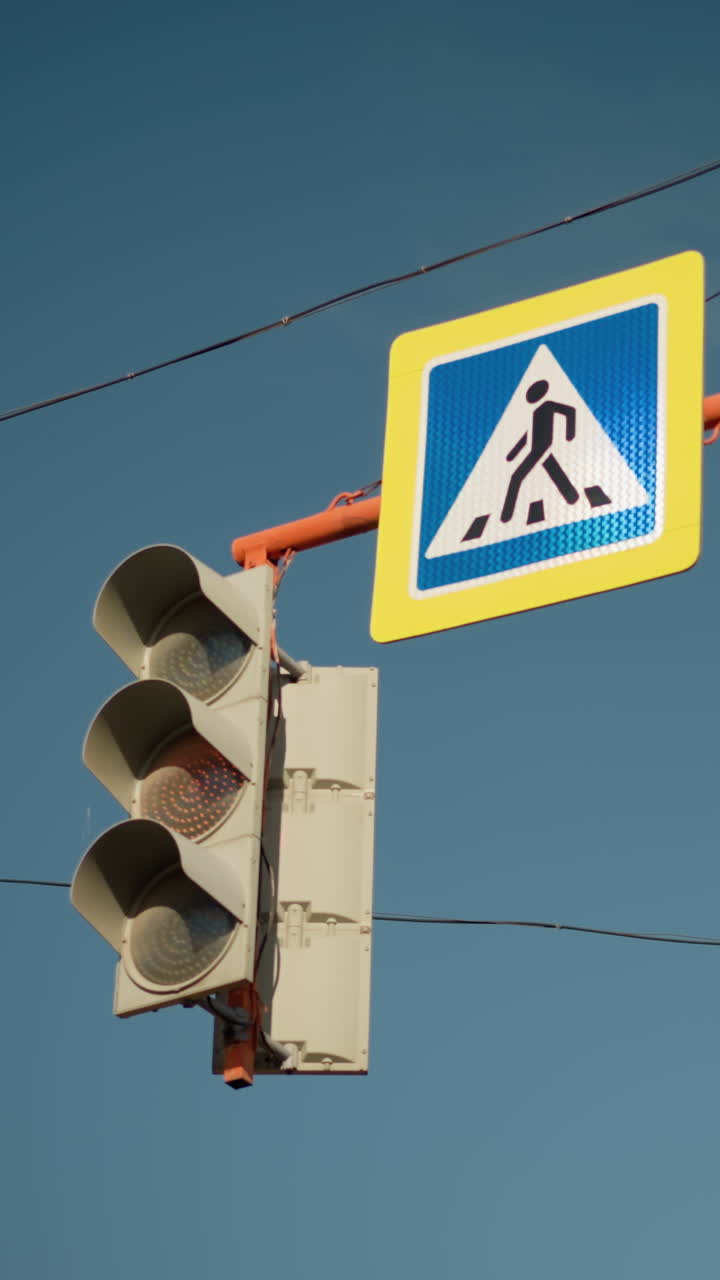 Semáforo y señal de peatones sobre una intersección, luz verde iluminada debajo del icono de cruce con marco amarillo, cableado aéreo y cielo azul despejado, carcasa del controlador mecánico y soporte de metal