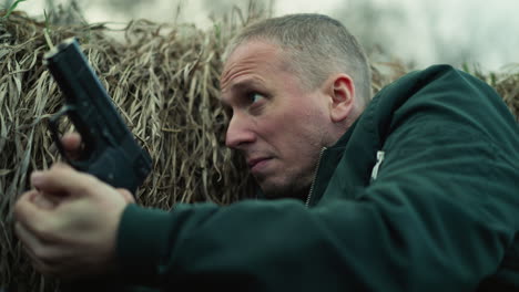 a close view of a man in a green jacket lying in a ditch against dry grass, aiming a pistol with intense focus