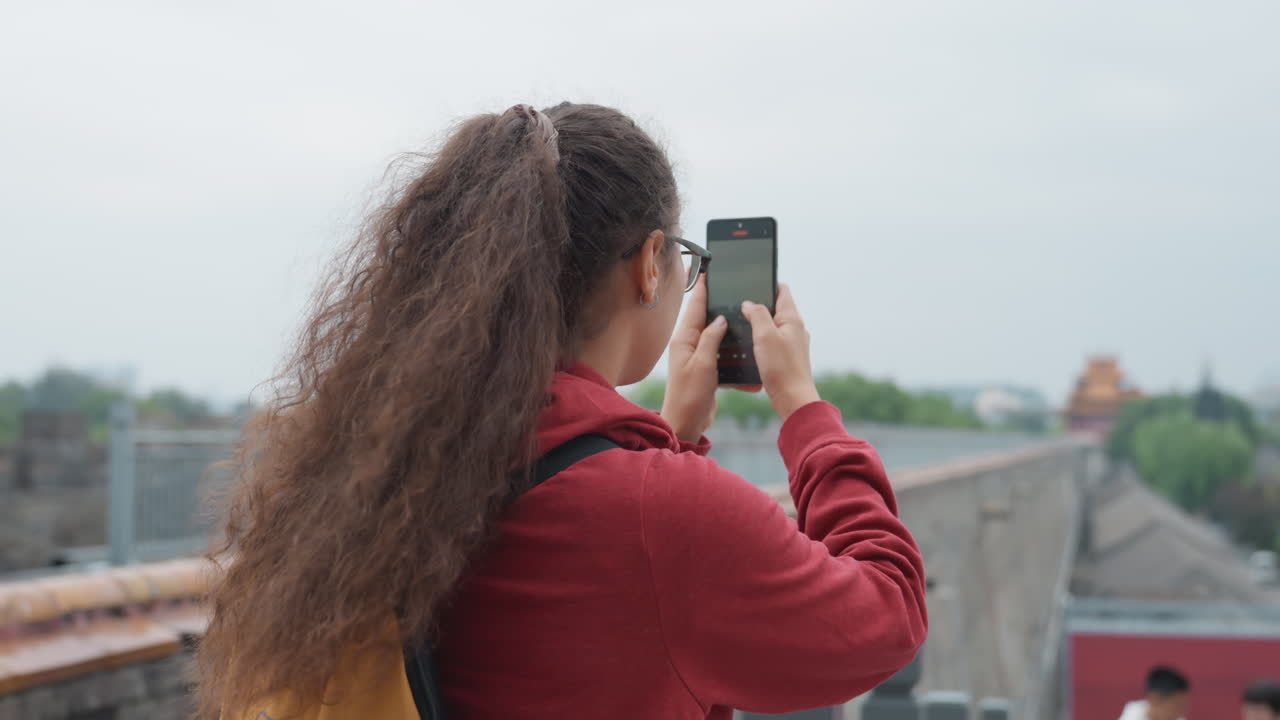 Jovem mulher fotografando o horizonte da cidade do telhado, cabelo cacheado em cascata, jaqueta vermelha, mochila amarela, smartphone segurado firmemente, céu nublado, marco distante além do corrimão de pedra, urbana calma.