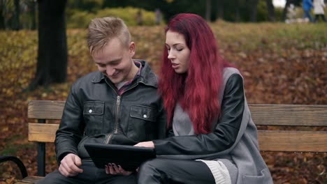 young woman with red hair and attractive man in a leather jacket sitting on a bench in park and using a digital talbe while discussing someting. they are choosing a trip and shopping online