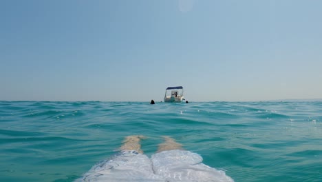 first-person pov of man legs relaxing and sunbathing while floating on sea water surface with boat in background