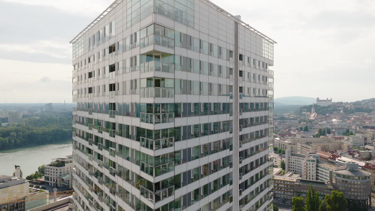 Drone shot of a tall residential building with glass balconies in Bratislava, Slovakia, overlooking the Danube River and Bratislava Castle in the distance