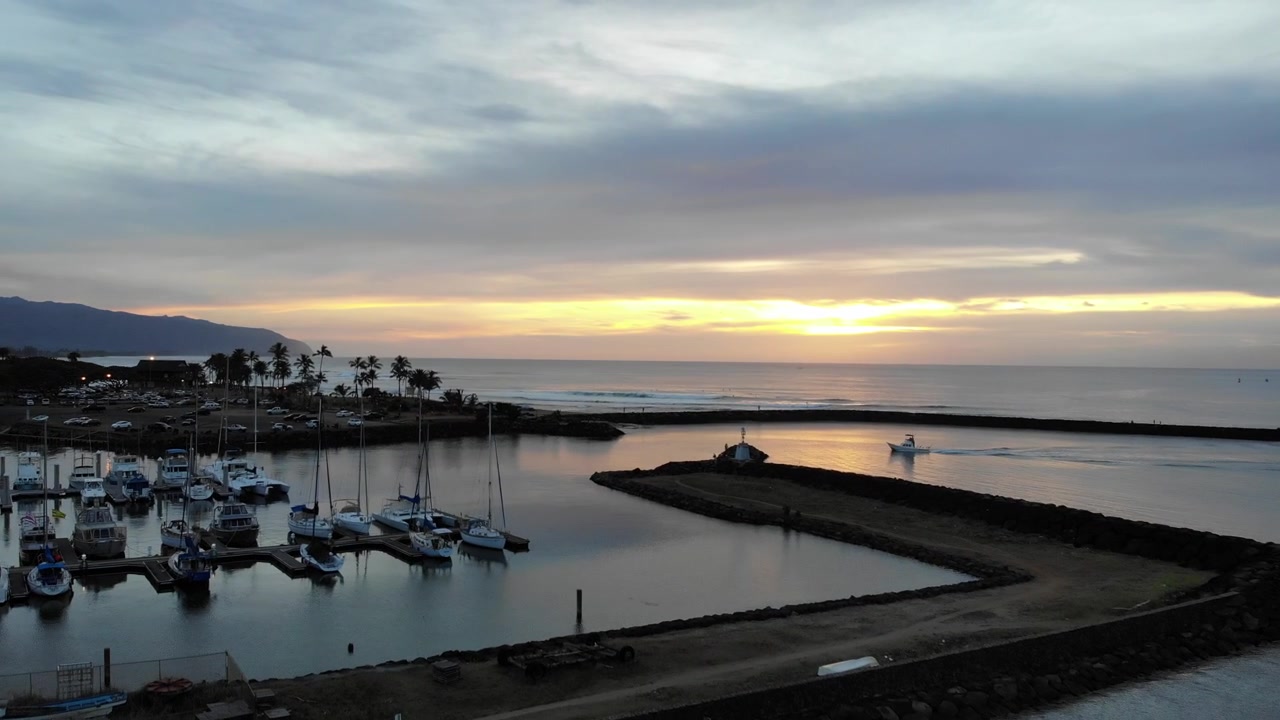 Toma de dron ascendente de una lancha motora entrando al puerto en Hawái, Haliva, durante un hermoso atardecer. También se ven palmeras y montañas al fondo.