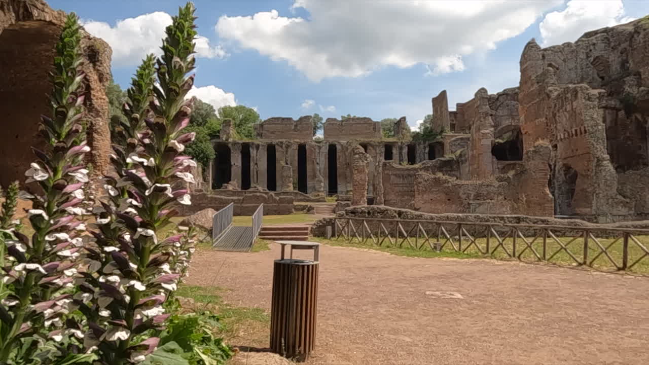 Antiche rovine romane a Villa Adriana a Tivoli vicino a Roma, Italia. Archi e mura storiche in pietra con piante fiorite bianche in primo piano sotto un cielo azzurro.