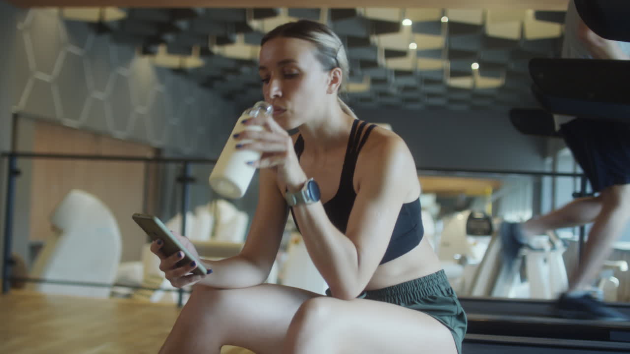 Woman Drinking Water and Using Phone in Gym