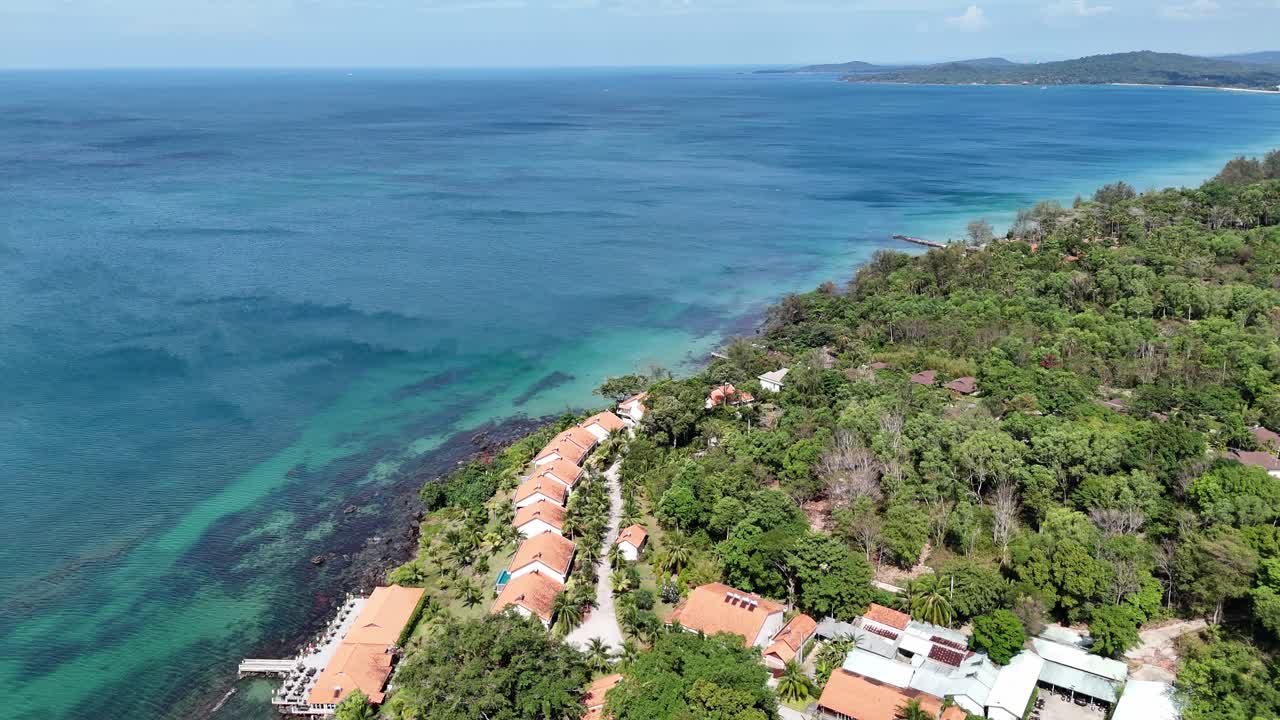 A coastal resort nestled along a pristine coastline. The resort boasts a cluster of white buildings with red roofs, blending with the lush green surroundings. A small pier extends blue waters