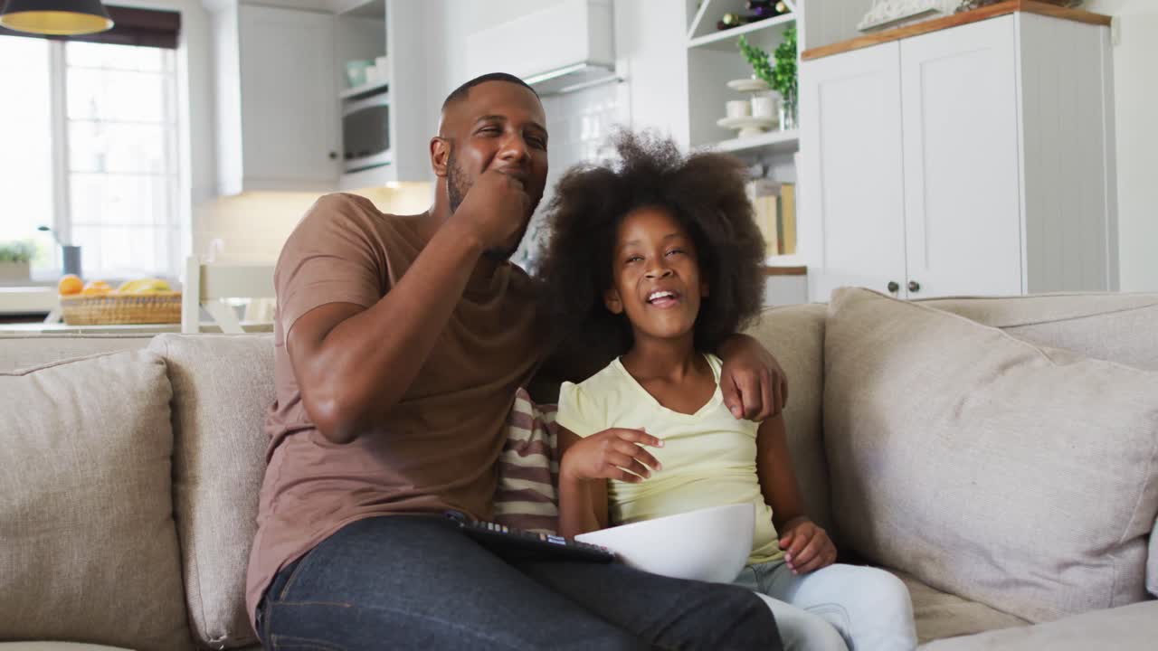 Figlia afroamericana e suo padre sul divano che mangiano popcorn e ridono guardando la TV.