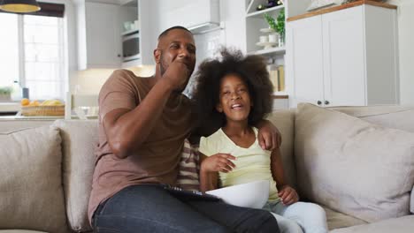 african american daughter and her father on couch eating popcorn and laughing at tv