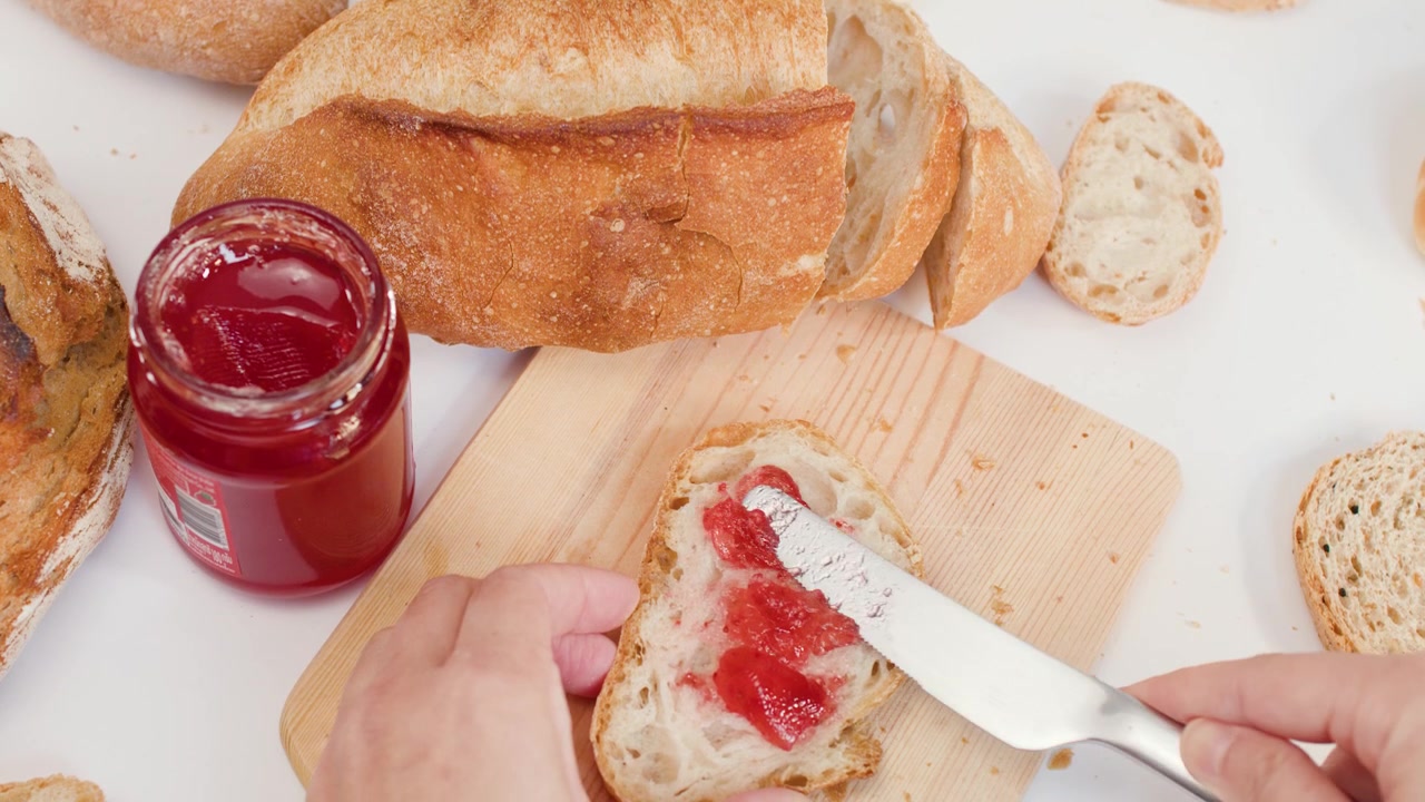 Top view of hands spreading red fruit jam on sourdough bread with a knife.