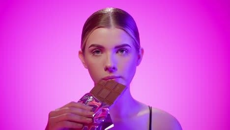 blonde nordic girl eats a chocolate bar suggestively in a studio with pink background, close up portrait shot