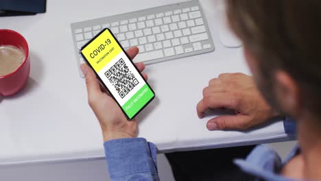 Mann-Am-Schreibtisch-Hält-Smartphone-Mit-Covid-Impfbescheinigung-Und-QR-Code-Auf-Dem-Bildschirm