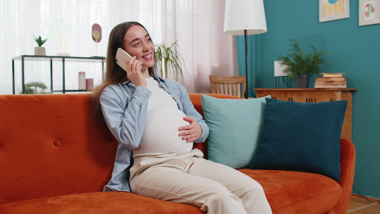 Smiling happy pregnant woman talking on smartphone stroking belly at home room pregnancy concept