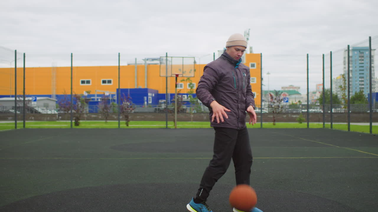 Atleta maschio che si allena a basket all'aperto, esercizio mattutino in solitaria su campo urbano con cielo nuvoloso, che porta una palla arancione, indossa una felpa con cappuccio e un berretto, esegue un tiro in sospensione, palleggio e movimento finale