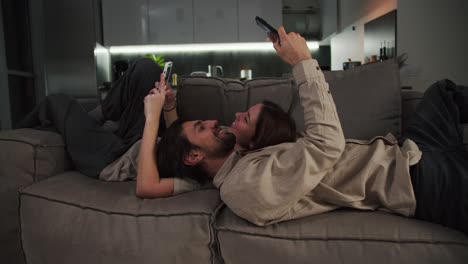 Happy-brunette-man-with-stubble-in-a-beige-shirt-lies-on-the-sofa-with-his-brunette-girlfriend-and-they-are-looking-at-something-on-their-smartphones-while-relaxing-on-the-sofa-in-a-modern-studio-apartment