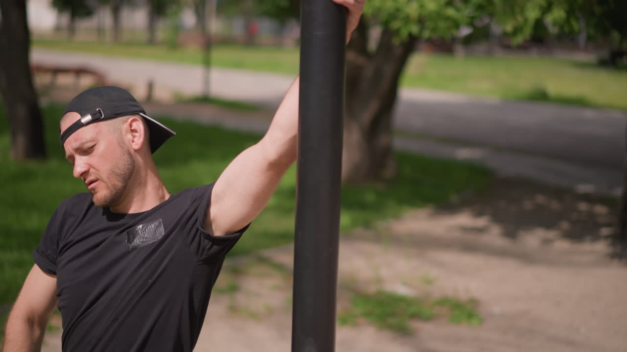 Wide Closeup Of Caucasian Man Leaning On Metal Pole In Park, Backward Cap, Black Shirt, Resting Posture, Soft Sunlight, Trees And Grass Background, Contemplative Urban Leisure Scene