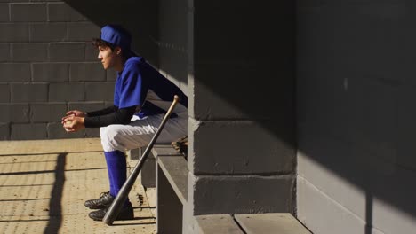 Nervöse-Gemischtrassige-Baseballspielerin,-Die-Auf-Einer-Bank-In-Der-Sonne-Sitzt-Und-Mit-Einem-Baseballball-Wartet