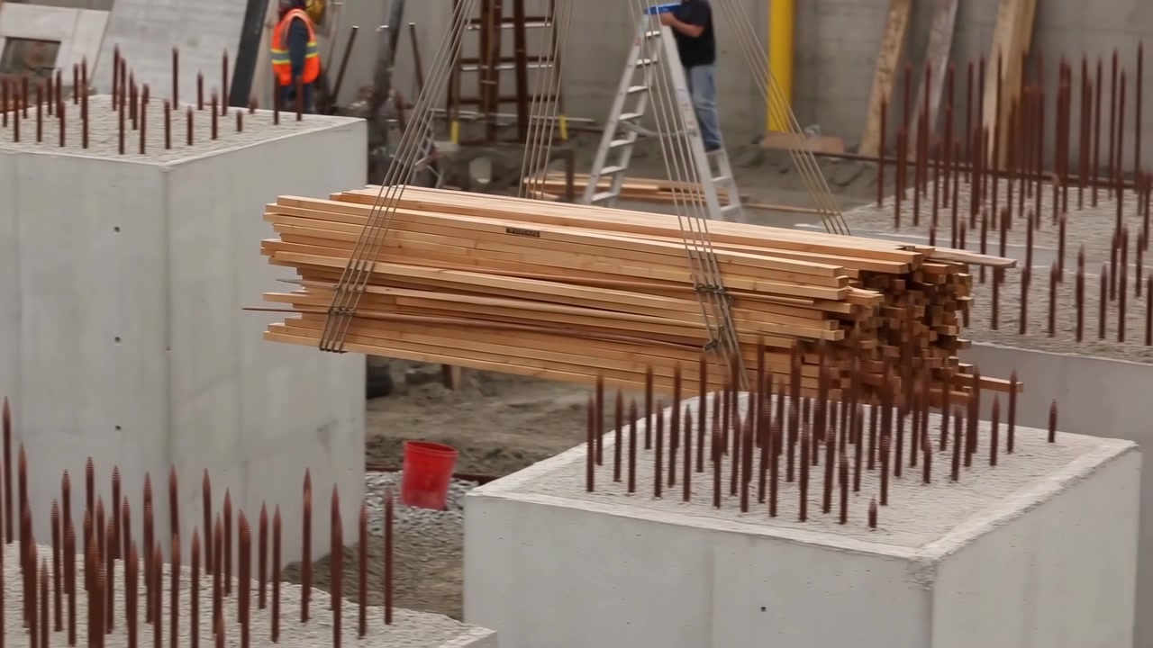 Fascio di legname piallato legato con cinghie metalliche pende da cavi d'acciaio sopra celle di fondazione in cemento colato, con fitte armature in ferro arrugginito che si ergono nelle vicinanze e la squadra del cantiere che ne guida il posizionamento attento.