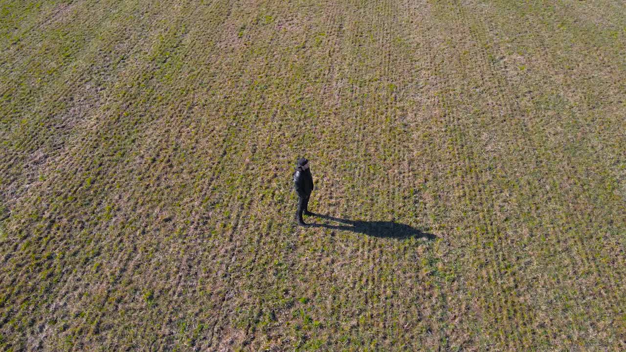 Aerial drone footage orbiting and spinning around a man with black clothing and a hat who is standing alone and still in a green and brown farmland or field during a sunny spring day, creating shadow.