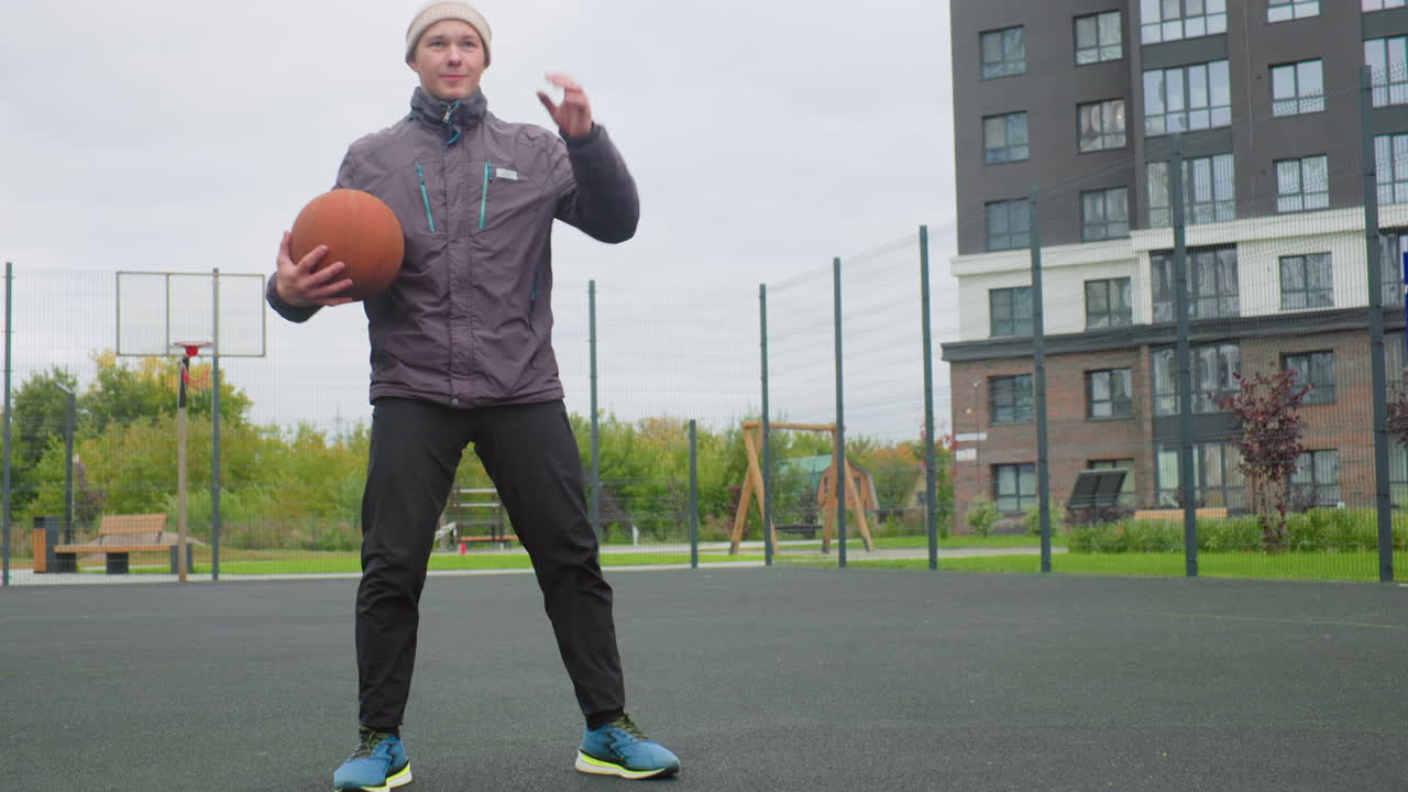 Hvid mand drible basketball på banen, laver crossover og boldhåndteringsøvelser, iført hue og jakke, sneakers banker mod asfalten i indhegnet park, lejlighedsbygning i baggrunden, skyet himmel