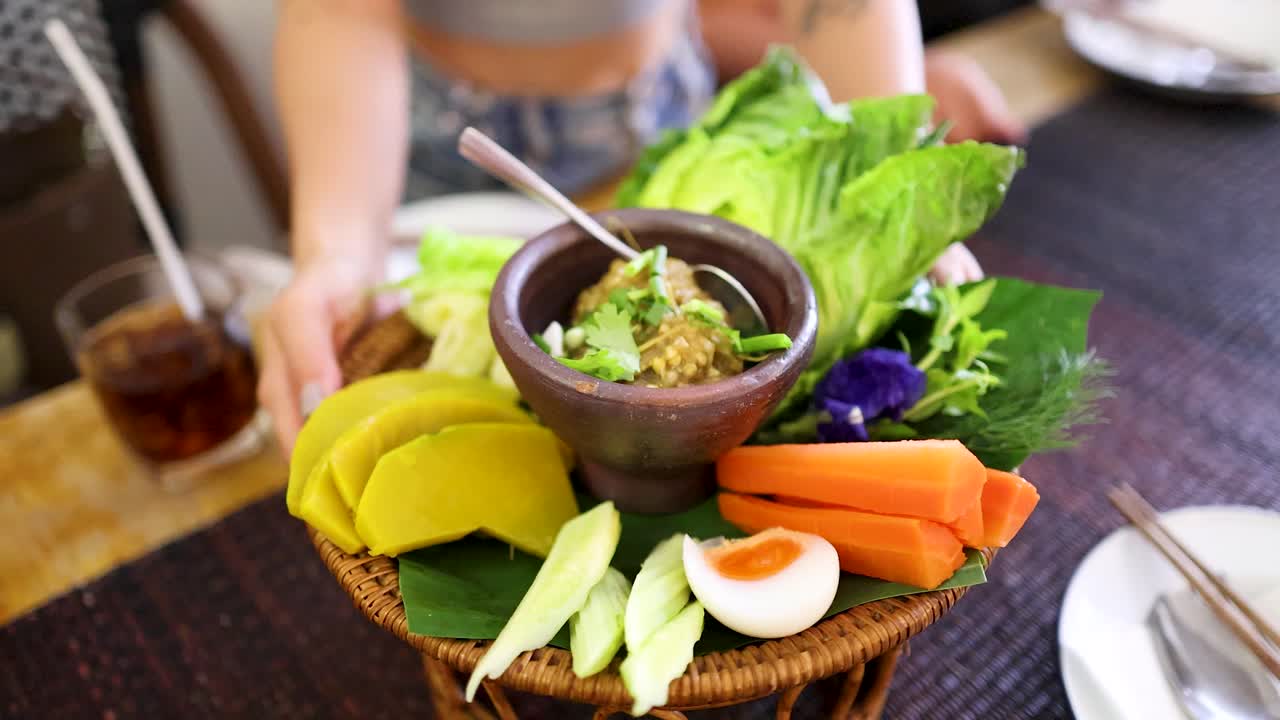 A person places a bamboo basket of Northern Thai roasted green chili dip, fresh vegetables, and boiled egg onto a dining table in bright, natural light