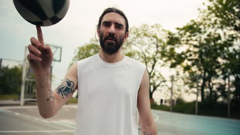 ritratto di un uomo serio in una maglietta bianca con una barba che gira una palla da basket sul suo dito indice sul campo da basket