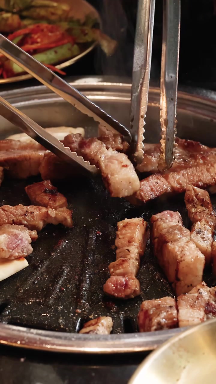 Potongan perut babi dipanggang di atas panggangan meja, penjepit membalik daging, pencahayaan hangat, tampilan jarak dekat.