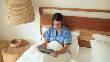 smiling woman in bed using tablet, wearing blue pajamas, enjoying morning, at home