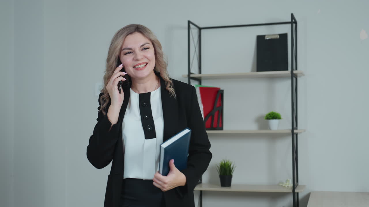 Cheerful professional woman speaking on phone, smiling with raised arm while holding blue-covered book, expressing excitement during conversation in bright office with shelves and decor in background