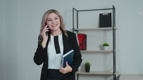 cheerful professional woman speaking on phone, smiling with raised arm while holding blue-covered book, expressing excitement during conversation in bright office with shelves and decor in background
