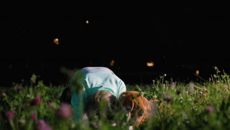 instructor kneels on grassy mat placing both hands down as obedient puppy mimics movement, learning commands during calm evening park training session surrounded by wildflowers under sunset sky