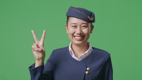 Close Up Of Asian Woman Air Hostess Smiling And Showing Peace Gesture To Camera While Standing In The Green Screen Background Studio