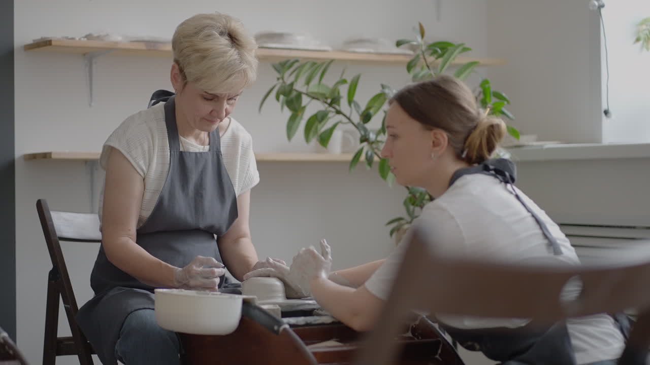 A female master shares her skills while giving a lesson for the elderly. Show grandmother the technique of working on a potter's wheel with ceramic clay