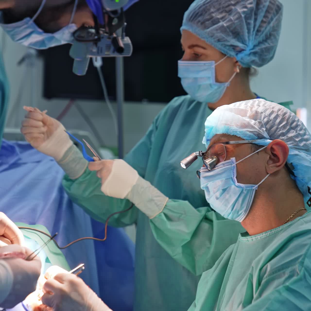 Chief neurosurgeon wearing device glasses working at surgery. Close up. Nurse at backdrop cleaning the instrument with sponge