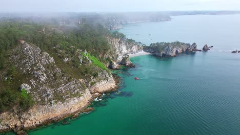 drone advancing above turquoise water toward île vierge beach on crozon peninsula, showing cliffs, rocks, morning mist and a small red boat - brittany in france