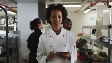 Portrait-of-african-american-female-chef-holding-tablet-looking-at-camera-and-smiling