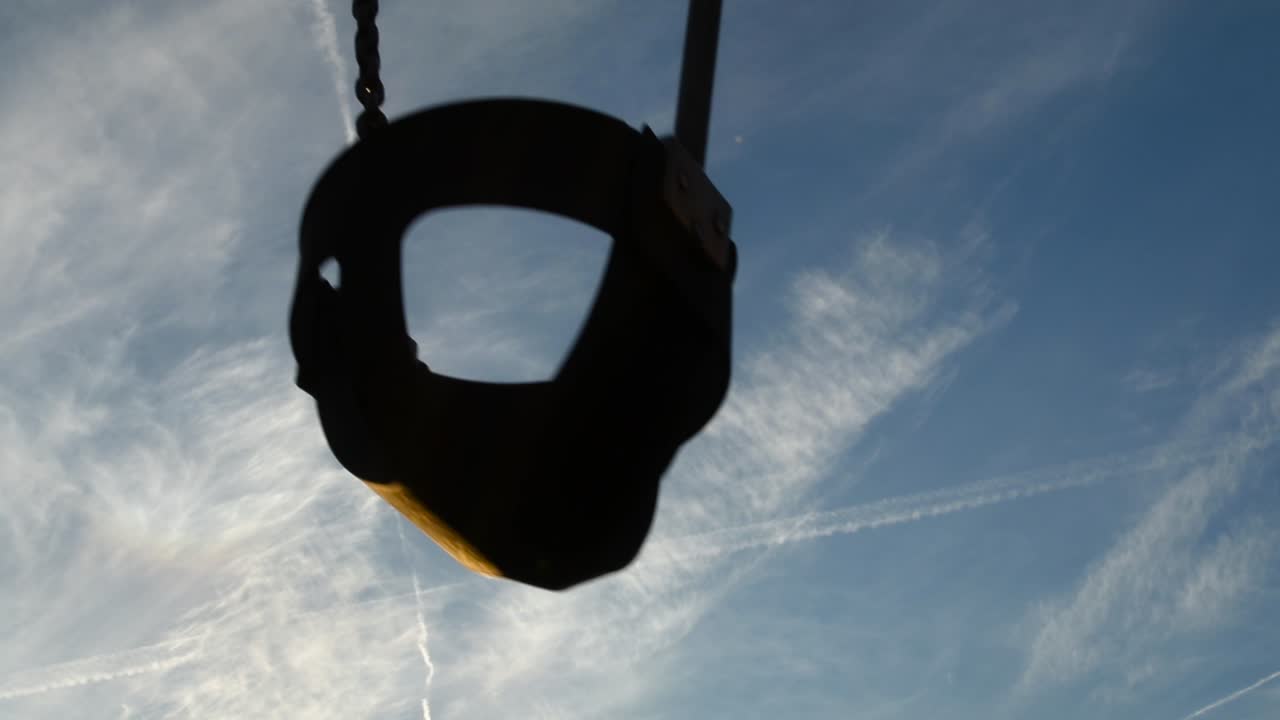 Empty children's swing rocks back and forth against blue cloud-filled sky. No kids on playground