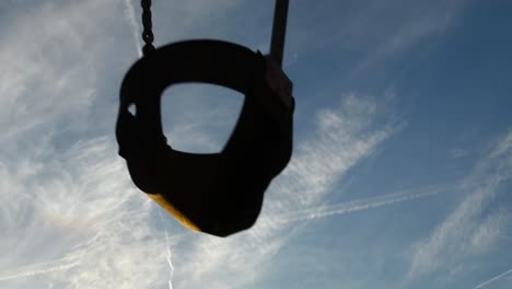 empty children's swing rocks back and forth against blue cloud-filled sky. no kids on playground